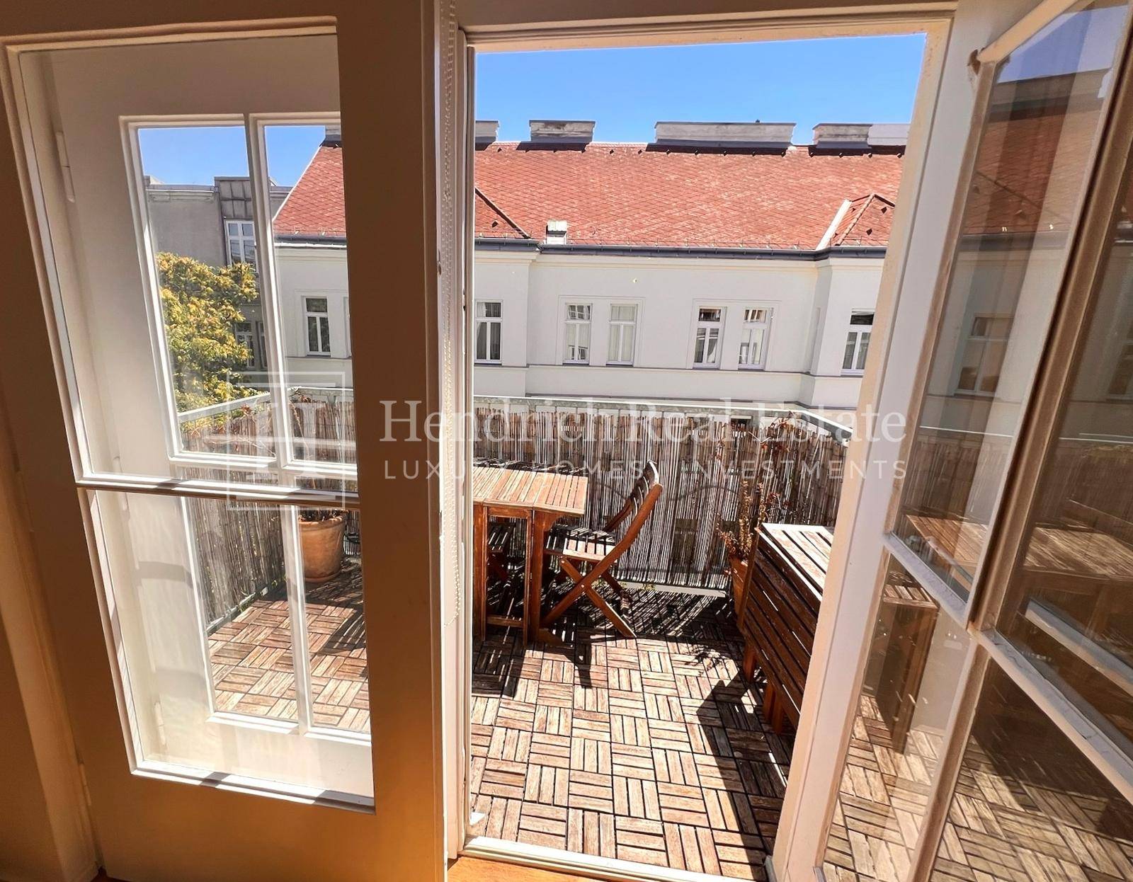 Blick aus dem Zimmer auf den sonnigen Balkon mit Holzboden und grüner Umgebung.