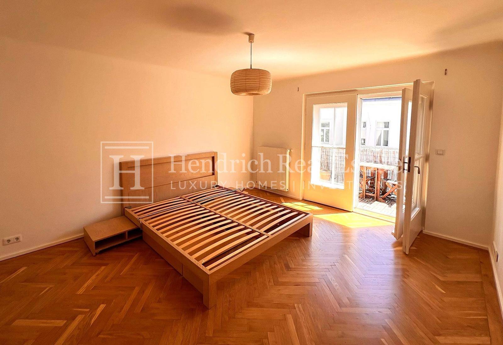Helles Schlafzimmer mit Fischgrätparkett, modernem Bettrahmen und Zugang zum Balkon.