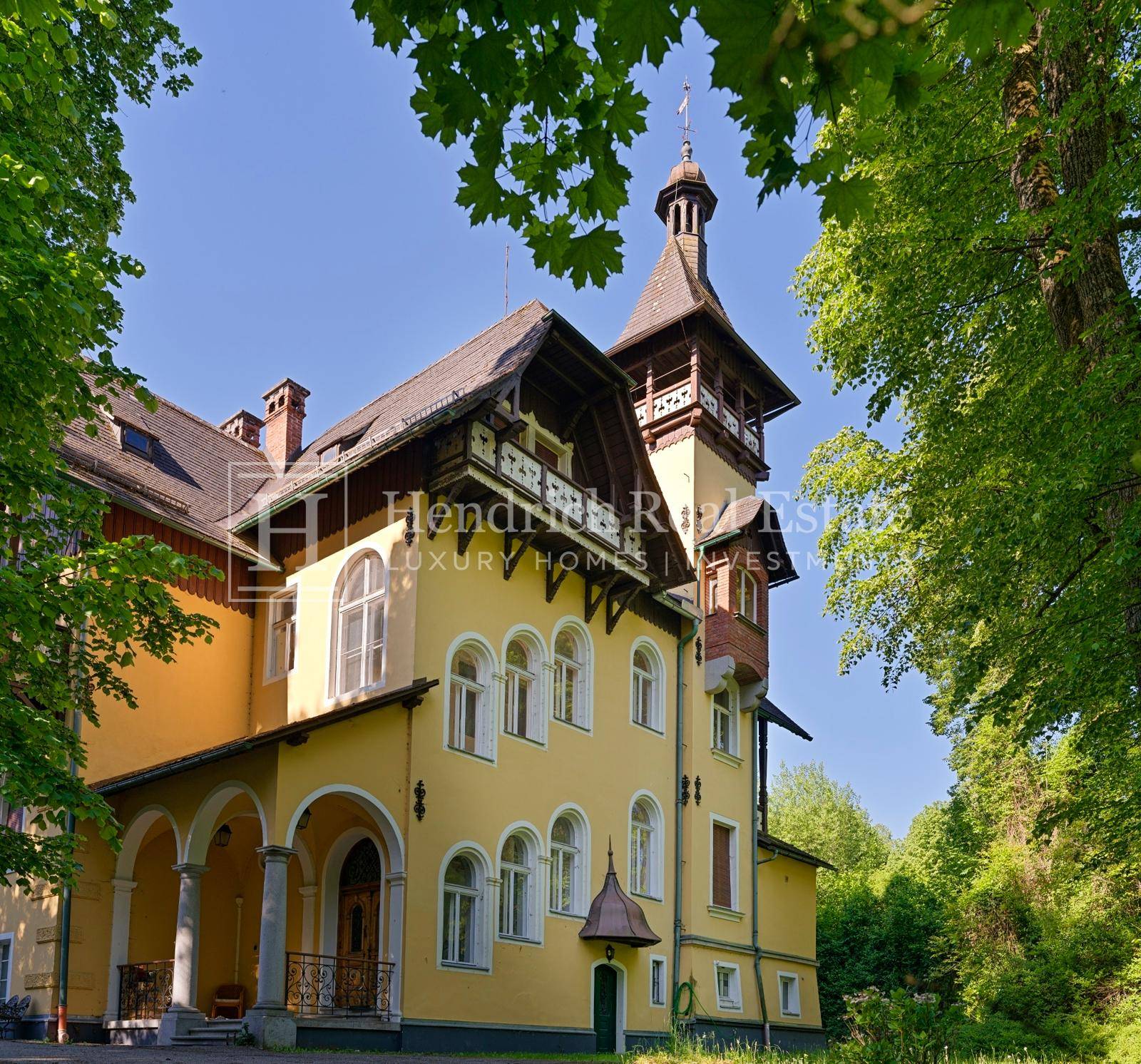 Historisches Gebäude mit markantem Turm und gelber Fassade, umgeben von üppigem Grün.