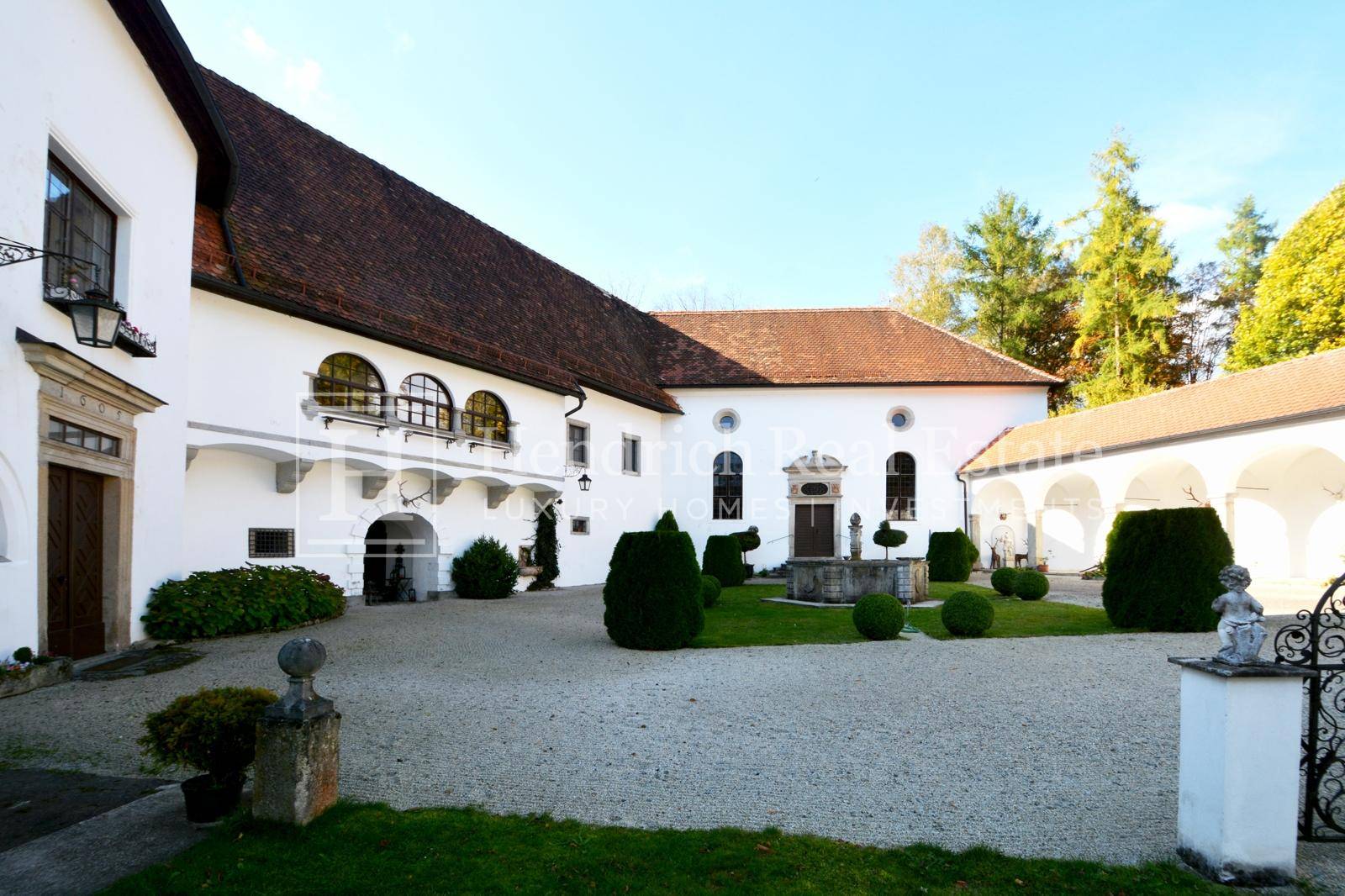 Historischer Innenhof mit Kiesfläche, gepflegten Büschen und einem alten Brunnen, umgeben von weißen Gebäuden.