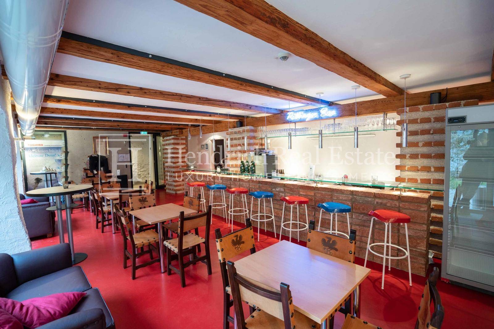 Gemütliche Bar mit Ziegeltheke, bunten Hockern und Holztischen unter sichtbaren Deckenbalken.