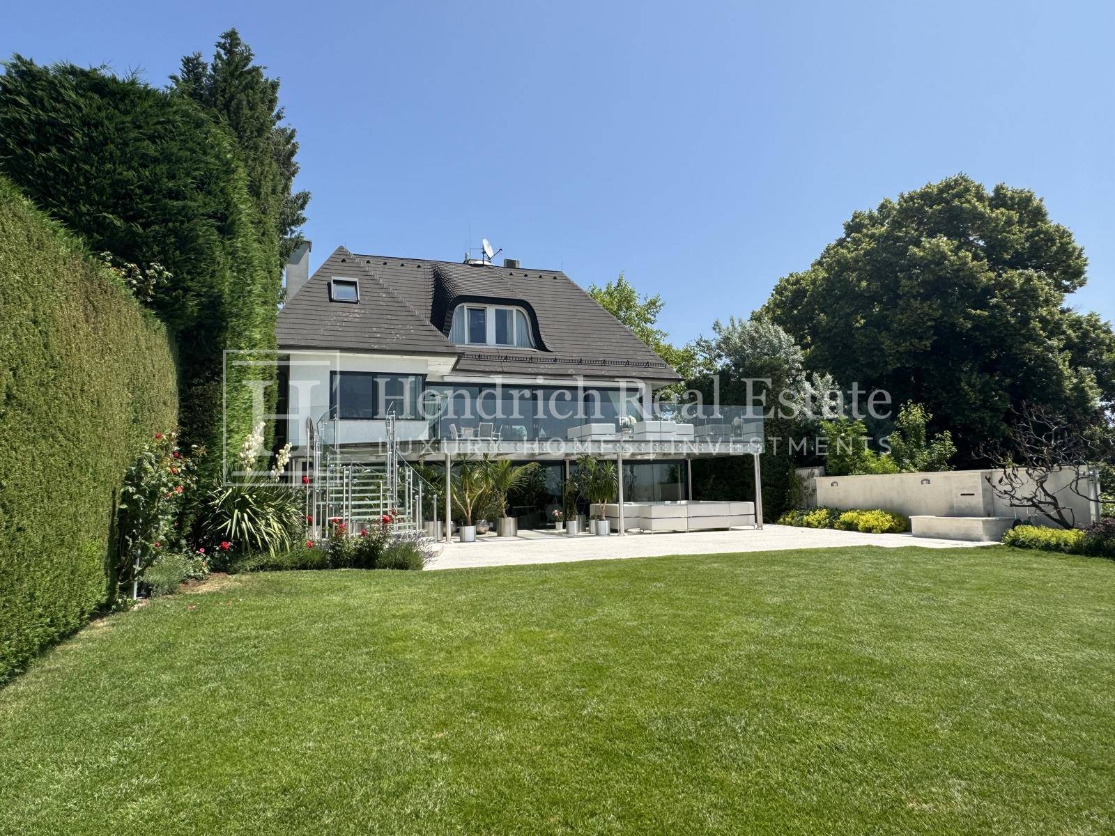 Moderne Villa mit großer Terrasse und weitläufigem Garten, umgeben von hohen Hecken und Bäumen.