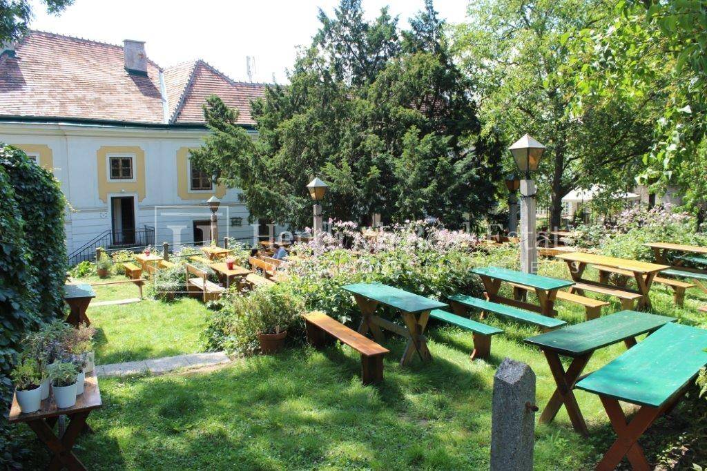 Idyllischer Gartenbereich mit zahlreichen Holztischen und Bänken unter schattenspendenden Bäumen.