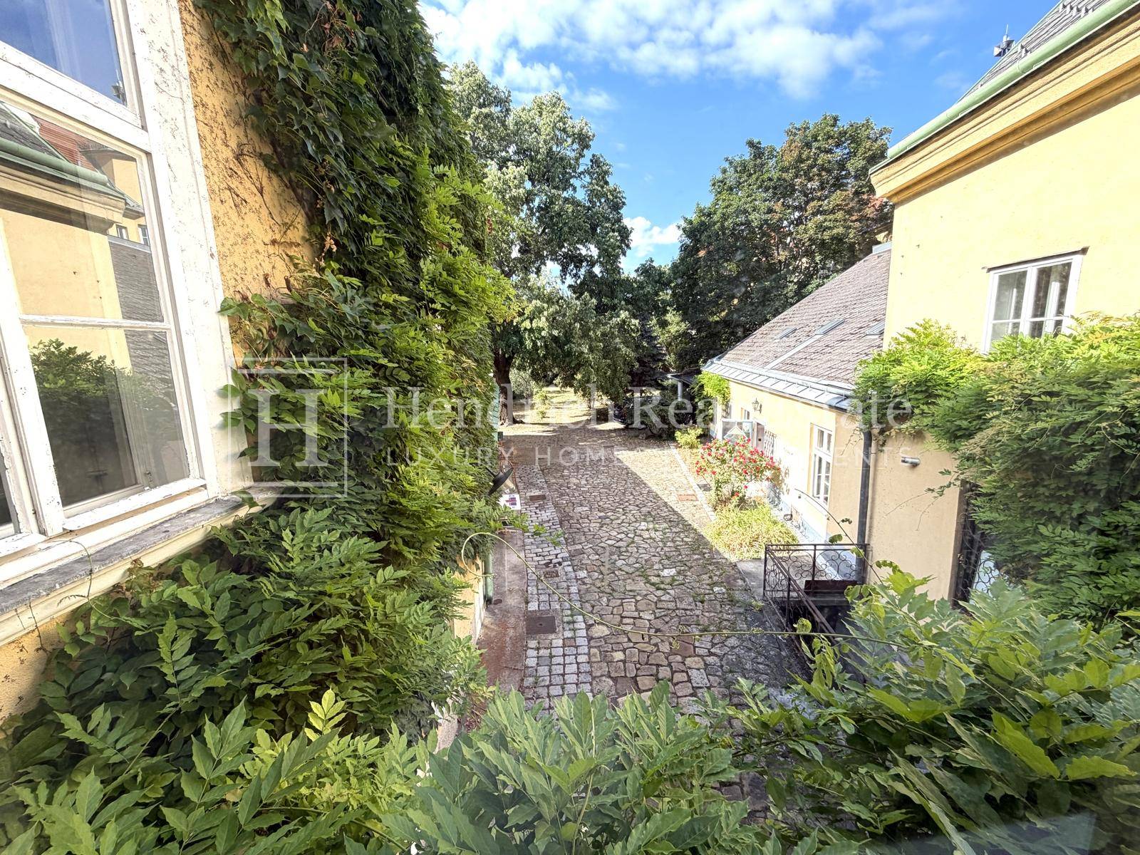 Gepflasterter Weg im grünen Gartenbereich, umgeben von üppiger Vegetation und alten Bäumen.