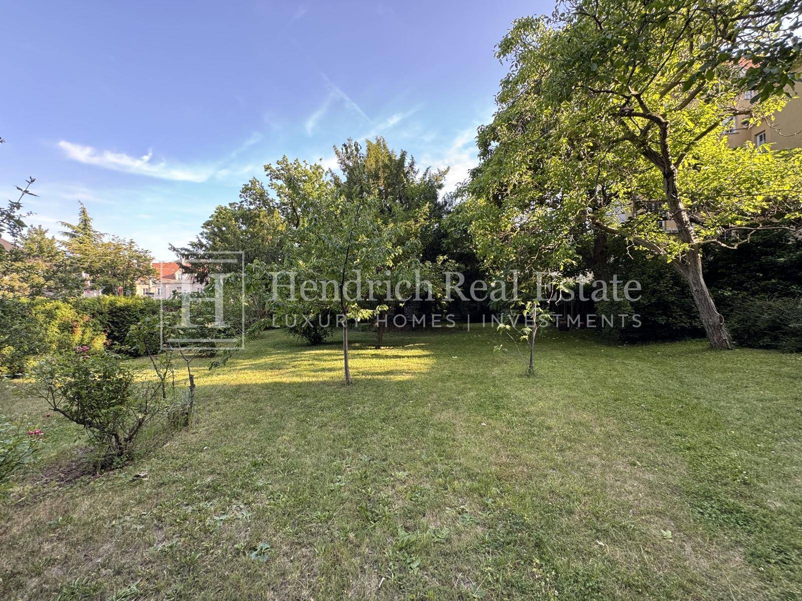 Weitläufiger Gartenbereich mit gepflegtem Rasen und altem Baumbestand unter blauem Himmel.