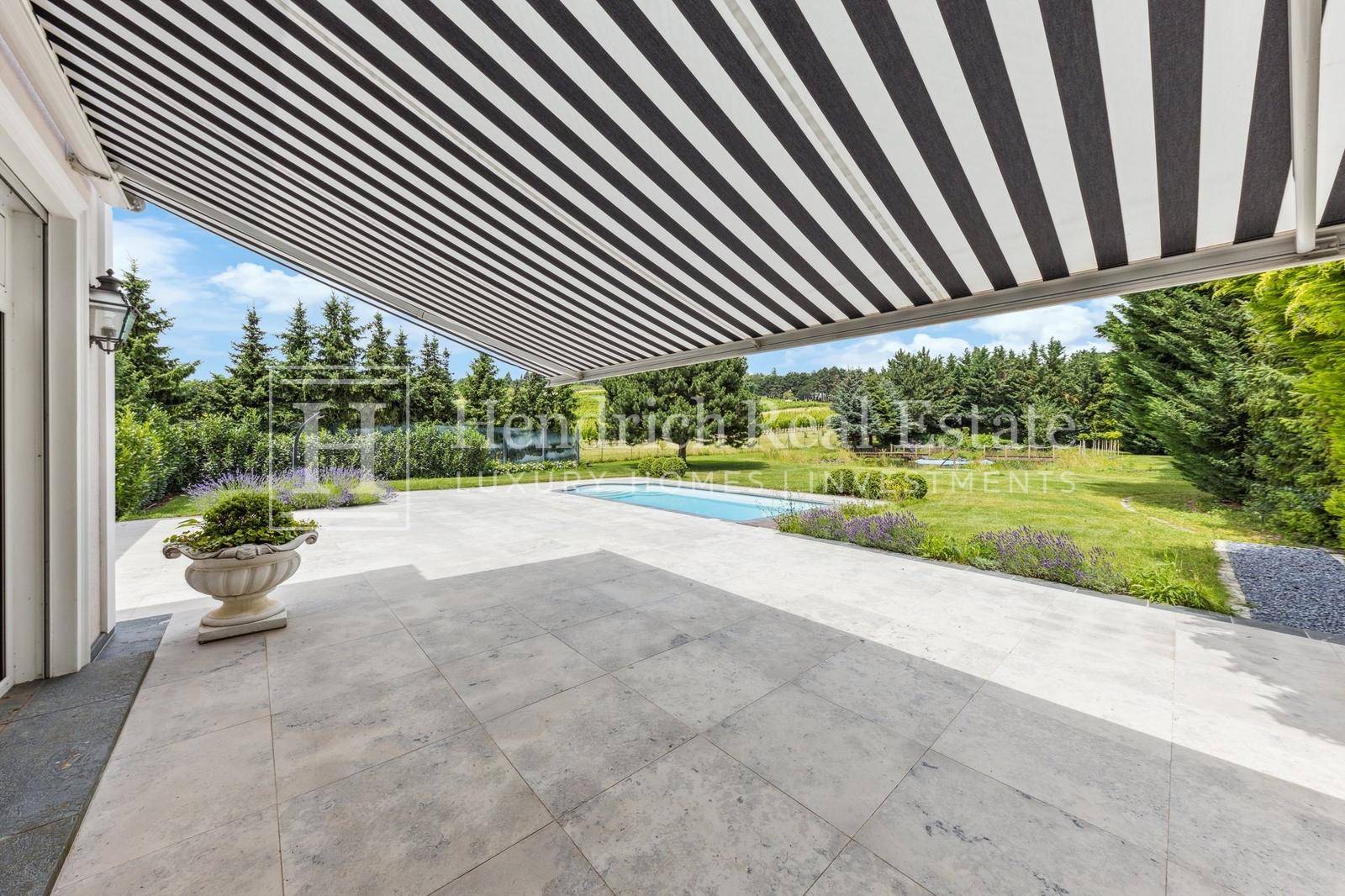 Weitläufige Terrasse mit gestreifter Markise, Blick auf den Pool und den gepflegten Garten.