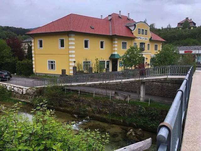 Gelbes Gebäude mit rotem Dach neben einem Fluss und einer Brücke in ländlicher Umgebung.
