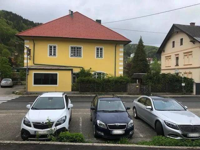 Gelbes Gebäude mit rotem Dach und parkenden Autos auf der Straße in ländlicher Umgebung.