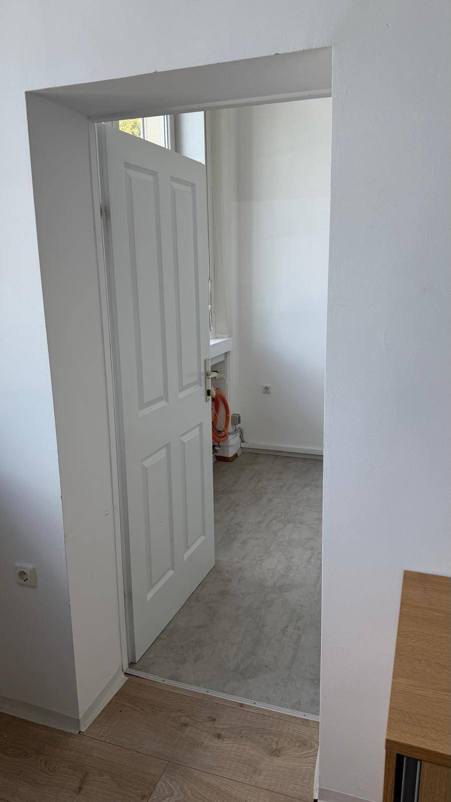 Türöffnung zu einem Raum mit grauem Boden, Fenster und Heizkörper, von einem Raum mit Laminatboden aus gesehen.