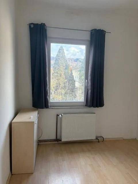 Leeres Schlafzimmer mit Holzboden, Fenster mit dunklen Vorhängen und Heizkörper.