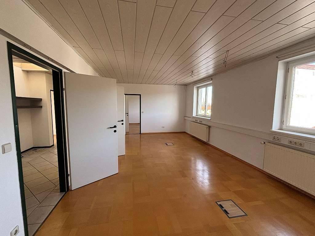 Geräumiger Büroraum mit hellem Holzparkett, weißen Wänden und zwei Fenstern, die Tageslicht hereinlassen.
