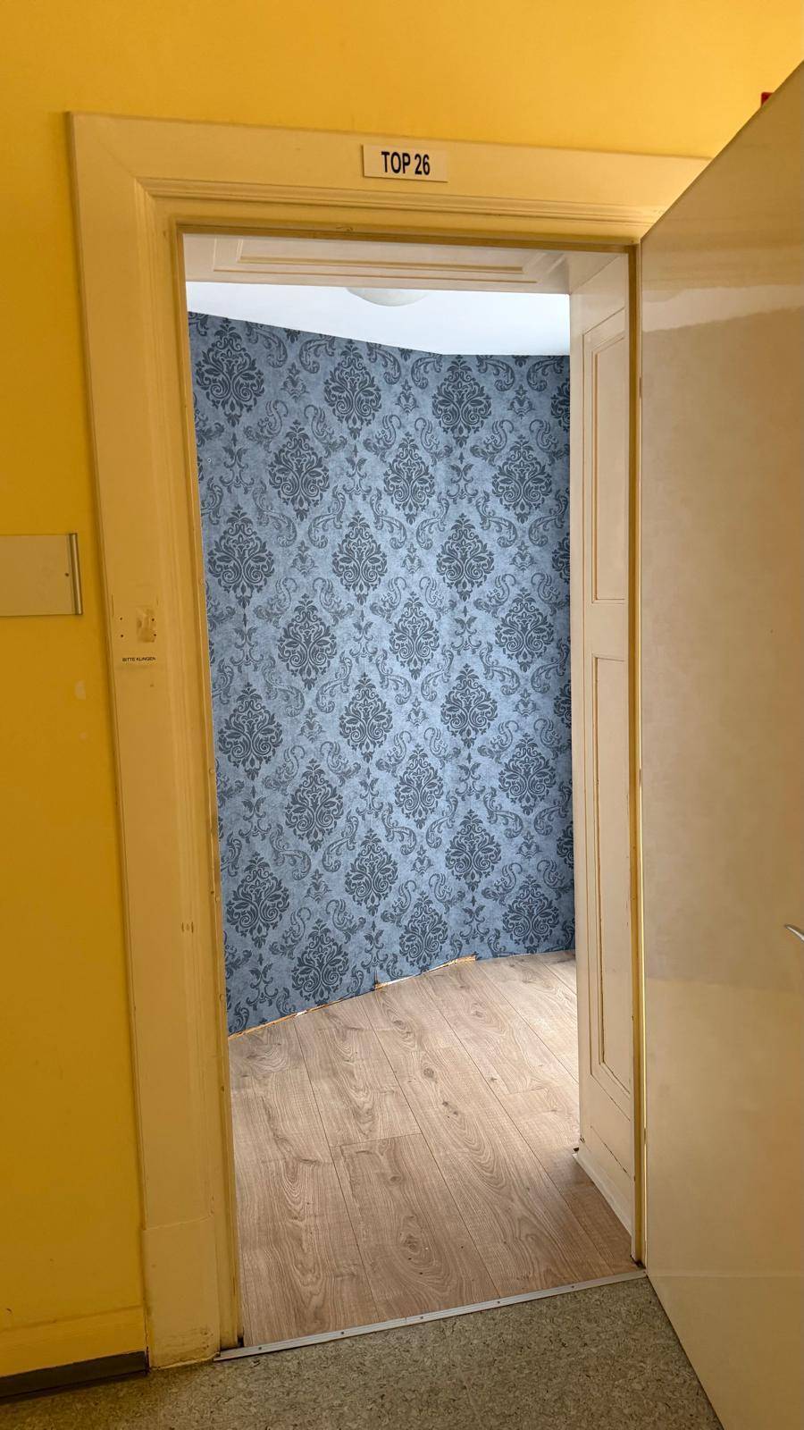 Eingangsbereich mit gelben Wänden und Blick in einen Raum mit gemusterter Tapete und Laminatboden.