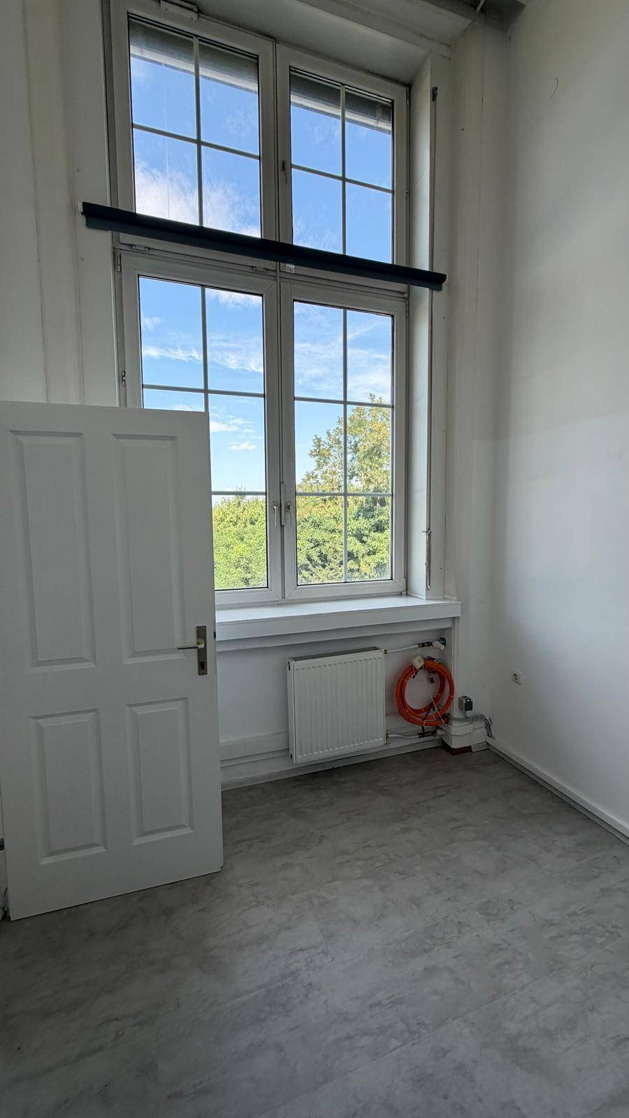 Kleiner Raum mit grauem Boden, großem Fenster, Heizkörper und orangefarbenen Kabeln, wirkt funktional.