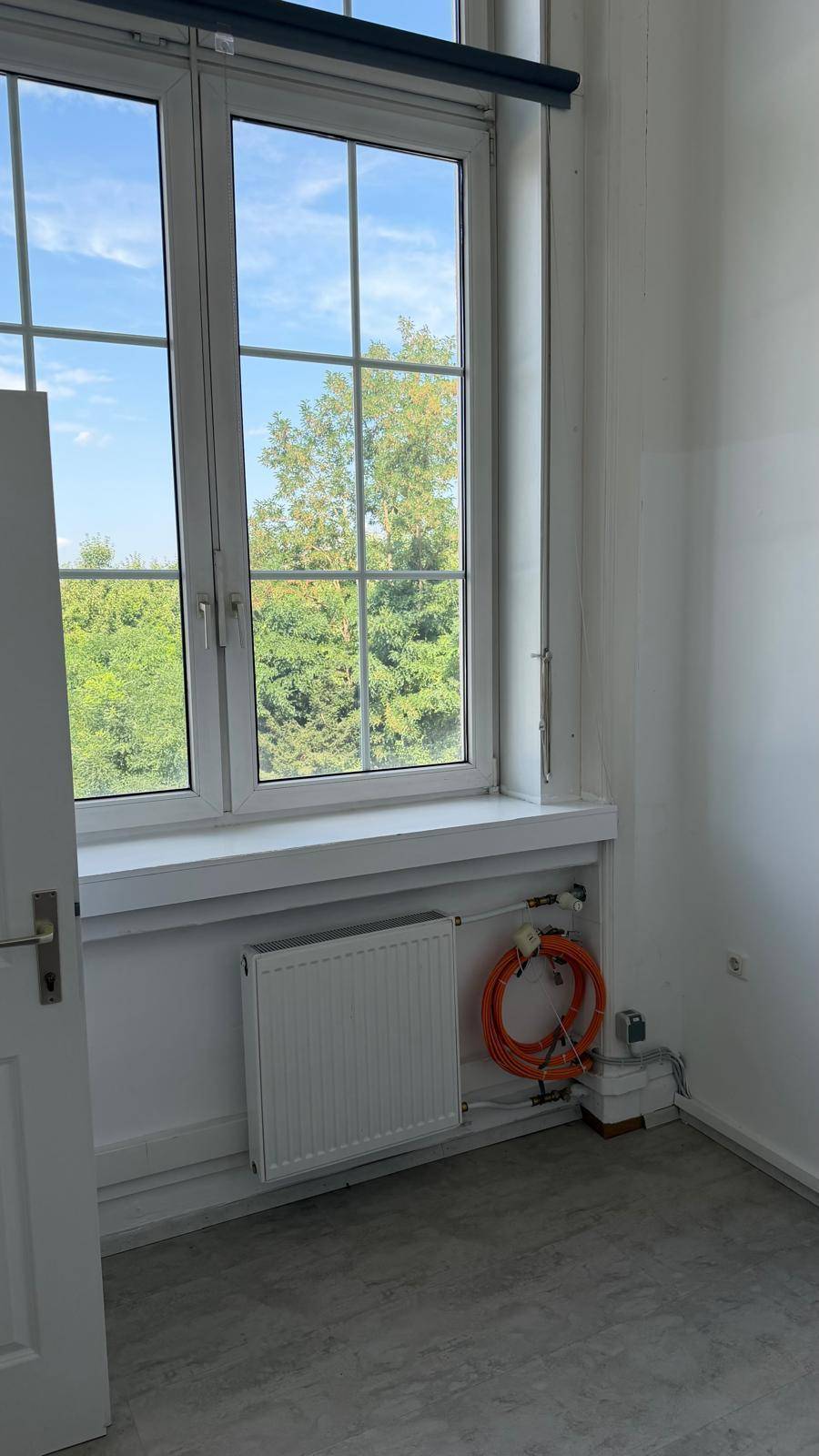 Kleiner Raum mit Fenster, Heizkörper und sichtbaren Kabeln auf dem grauen Boden, möglicherweise ein Abstellraum.
