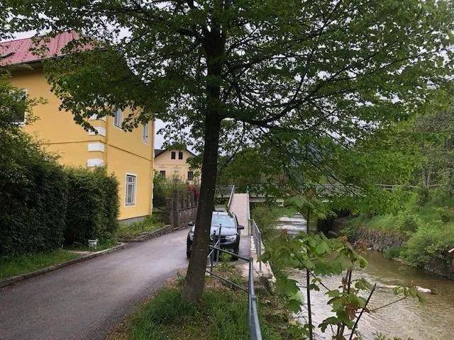 Straße entlang eines Flusses mit Bäumen und einem gelben Gebäude im Hintergrund.