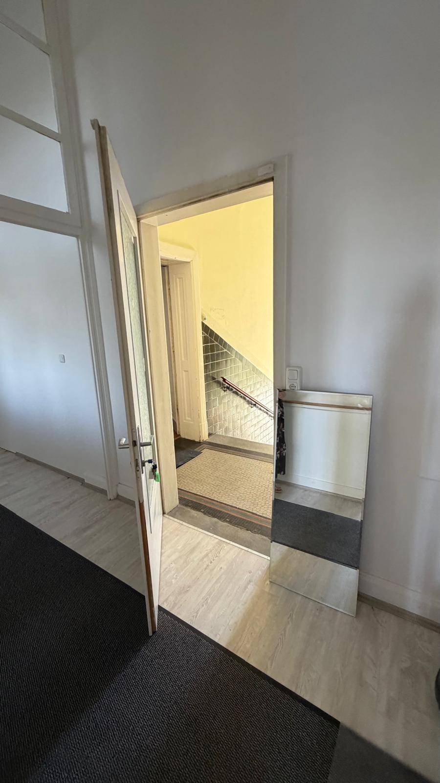 Flur mit Teppichläufer und offener Tür, die den Blick auf ein Treppenhaus mit gelben Wänden freigibt.