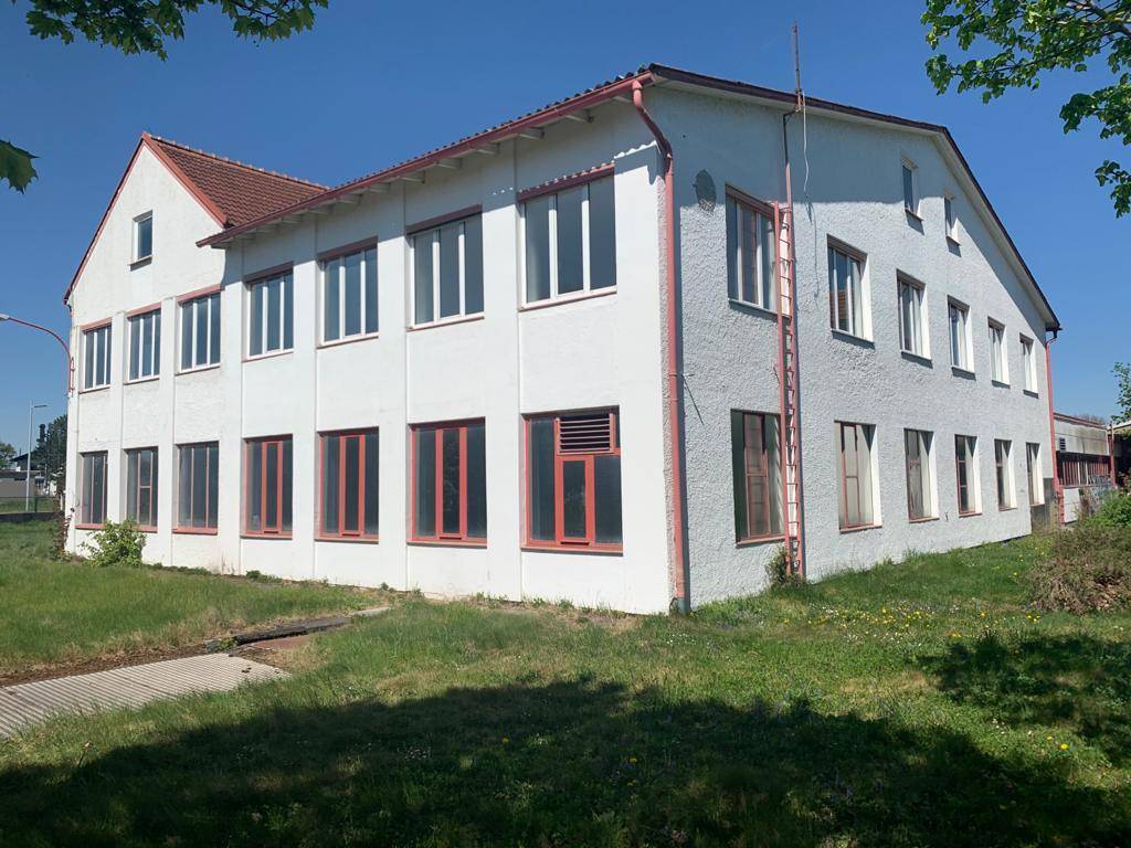 Weißes Gewerbegebäude mit roten Fensterrahmen und Dachkanten, umgeben von Grünflächen unter blauem Himmel.