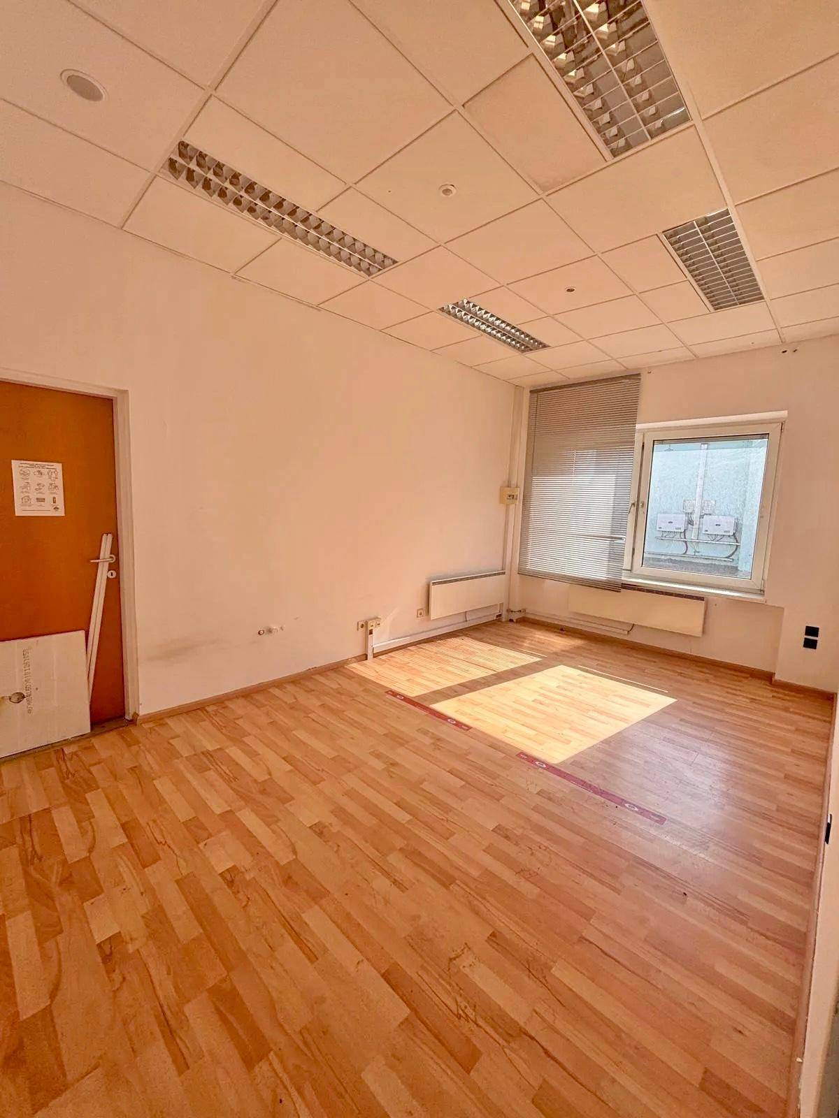 Leeres Büro mit hellem Holzboden, weißen Wänden und einem Fenster mit Jalousien, durch das Sonnenlicht fällt.