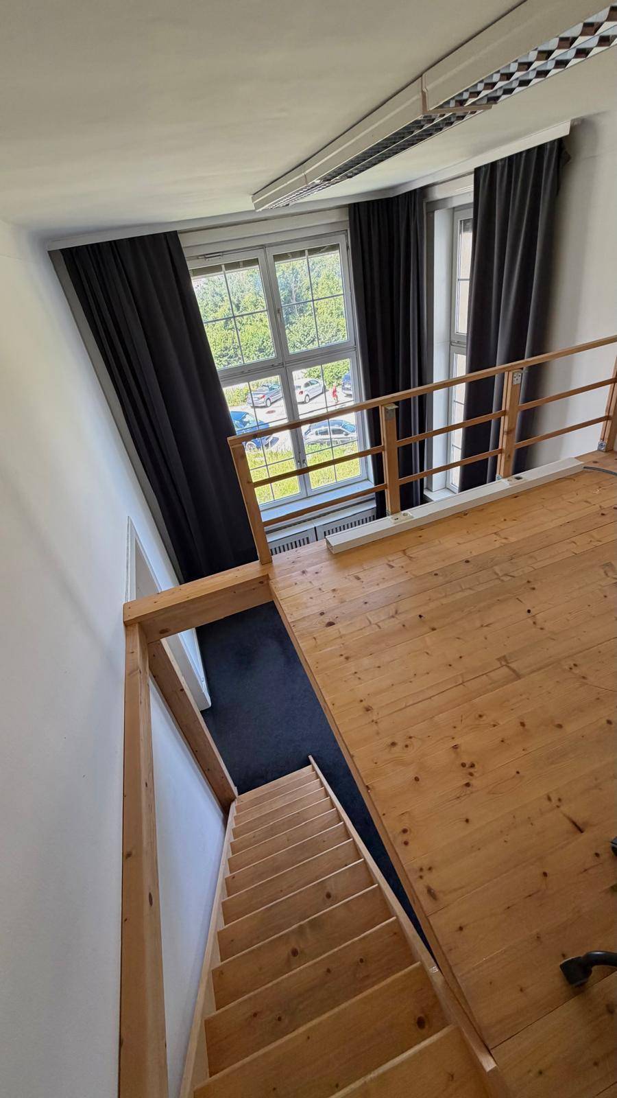 Blick von der Empore auf die Holztreppe und den unteren Bereich mit Fenstern und dunklen Vorhängen.