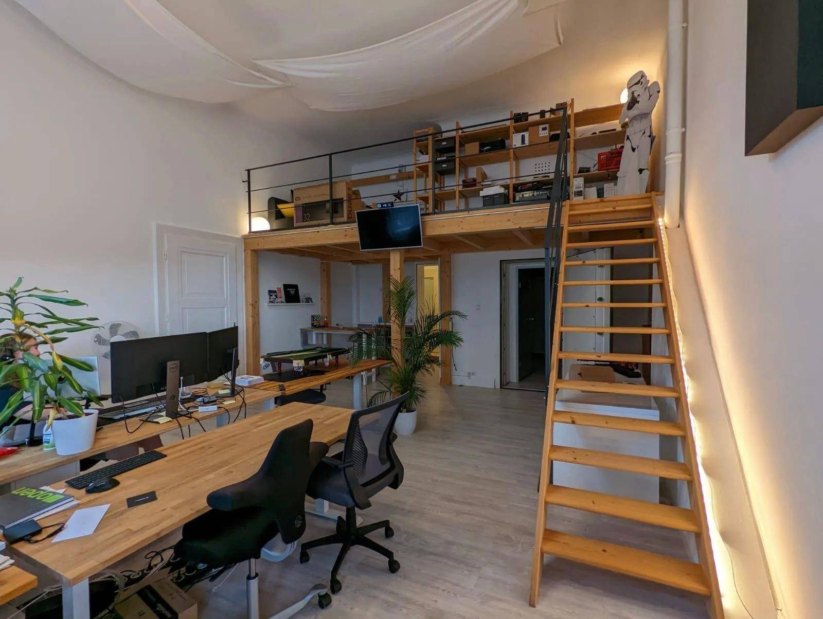 Großer Büroraum mit mehreren Arbeitsplätzen, Pflanzen und einer Holz-Loft-Ebene mit Regalen.