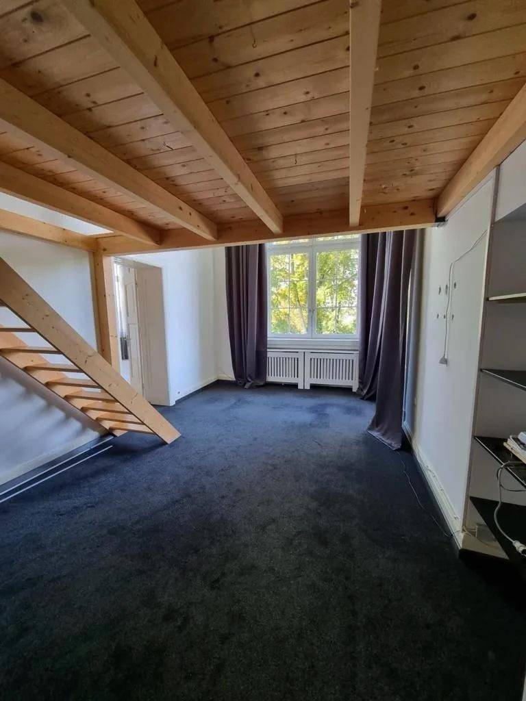 Gemütliches Zimmer mit dunklem Teppichboden, Fenster und einer Holz-Loft-Struktur mit sichtbaren Balken.