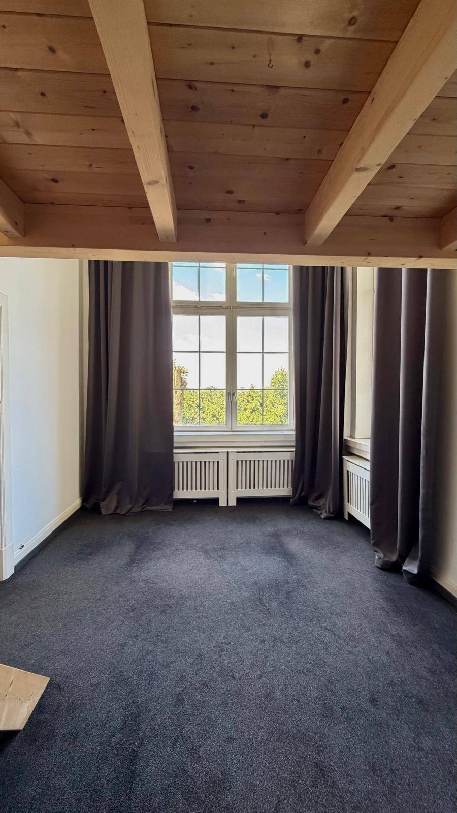 Wohnraum mit dunklem Teppichboden, sichtbaren Holzdeckenbalken und großen Fenstern mit Vorhängen.