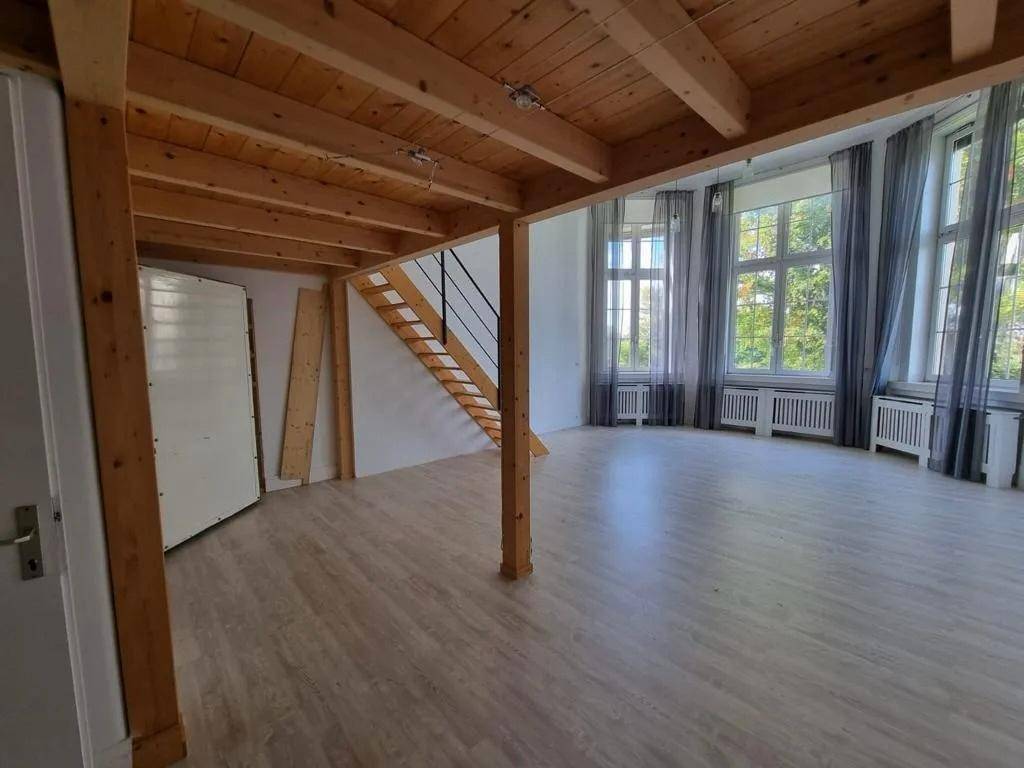 Heller, leerer Raum mit Holzbalkendecke, Laminatboden und großen Fenstern mit grauen Vorhängen.