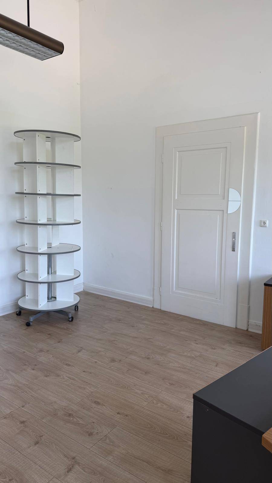 Geräumiges Büro mit Laminatboden, einer weißen Tür und einem runden Regal im Hintergrund, bietet vielseitige Nutzungsmöglichkeiten.