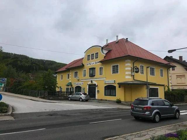 Gelbes Gebäude mit rotem Dach an einer Straße mit parkenden Autos und Hügeln im Hintergrund.