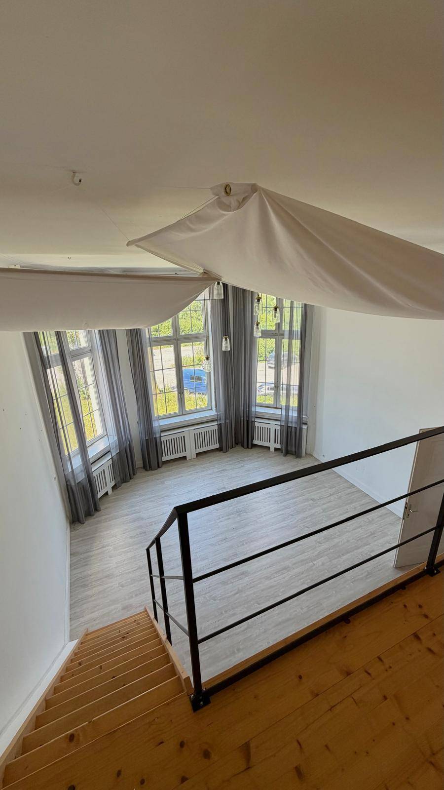 Blick von der Empore in den unteren Wohnbereich mit hohen Fenstern und hellem Laminatboden.