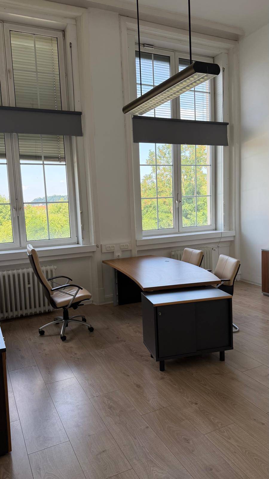 Helles Büro mit modernem Schreibtisch, Bürostühlen und hohen Fenstern mit Jalousien, die viel Tageslicht hereinlassen.