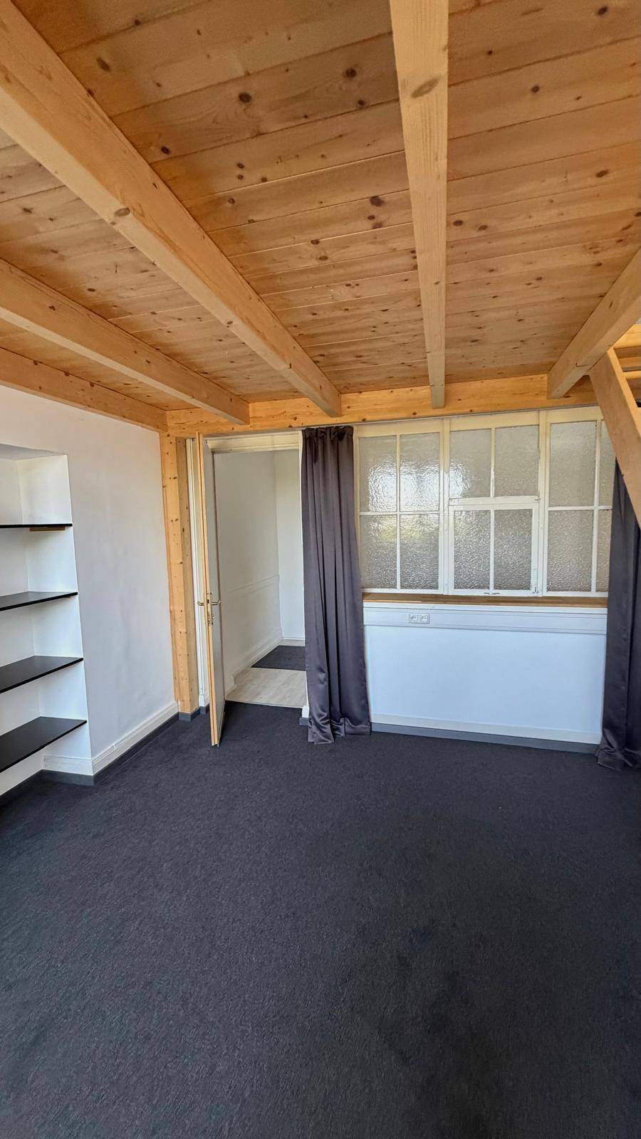 Wohnraum mit dunklem Teppichboden, sichtbaren Holzdeckenbalken und einem Fenster mit Sprossen.