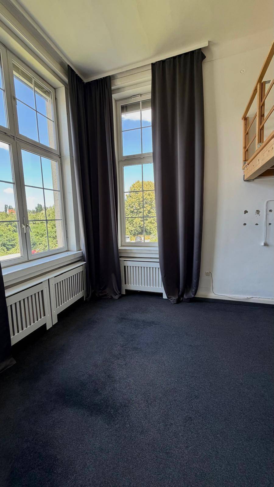Heller Wohnraum mit dunklem Teppichboden und großen Fenstern, die viel Tageslicht hereinlassen.