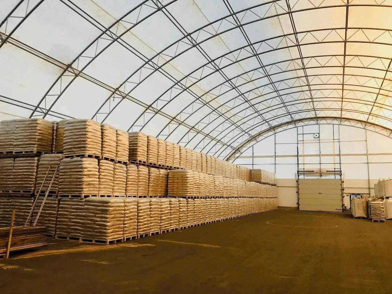 Innenansicht einer großen Lagerhalle mit gewölbtem Dach, gefüllt mit gestapelten Säcken und Metallkonstruktion.