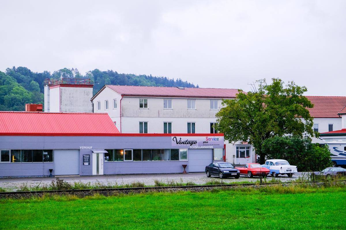 Gewerbegebäude mit rotem Dach und grauer Fassade, davor parken Autos auf einem Schotterplatz.