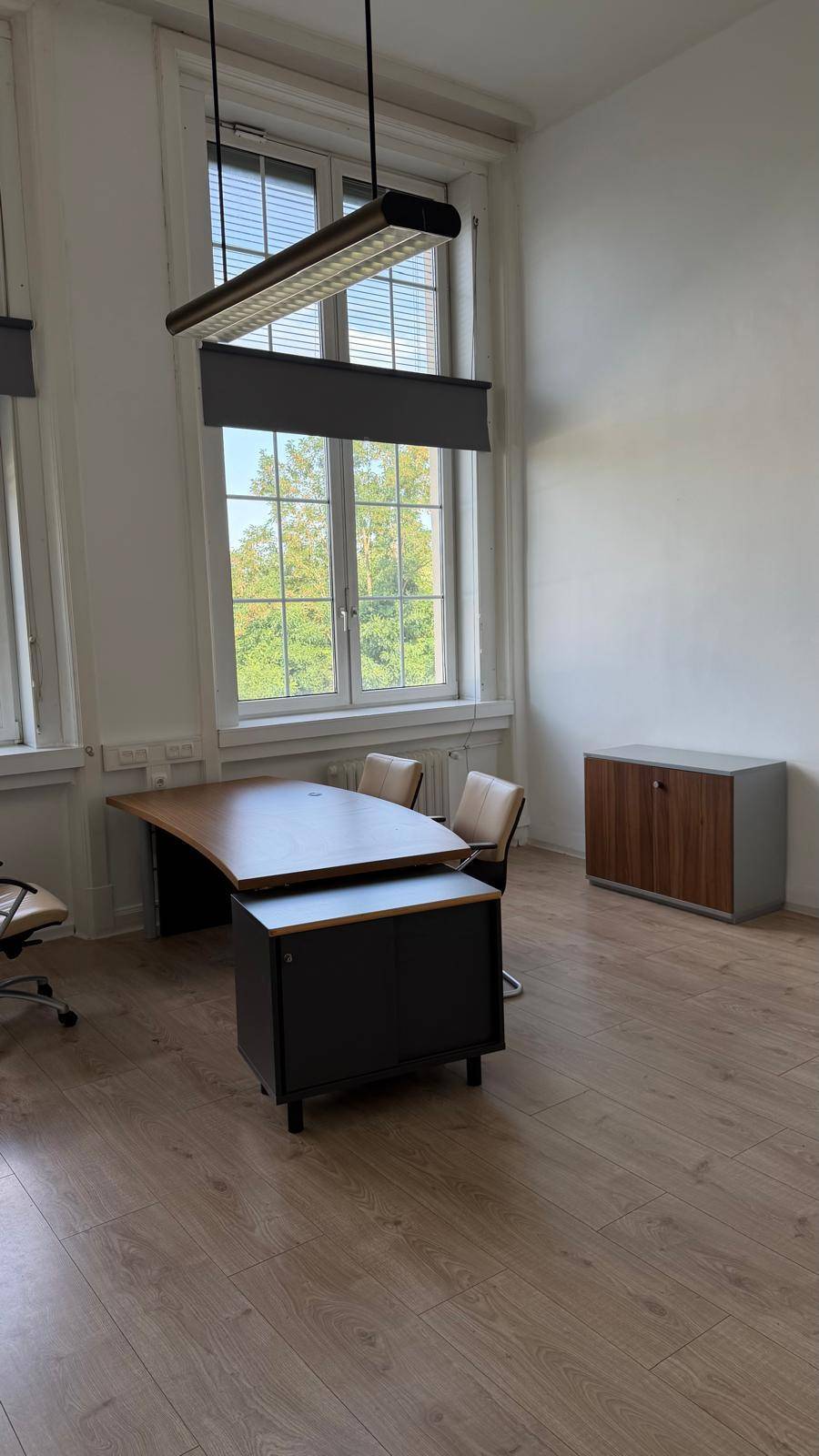 Helles Büro mit modernem Schreibtisch, Bürostühlen und hohen Fenstern, die den Raum mit viel Tageslicht versorgen.