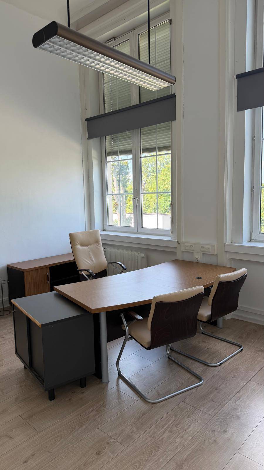 Helles Büro mit großem Schreibtisch, bequemen Stühlen und hohen Fenstern mit Rollos.