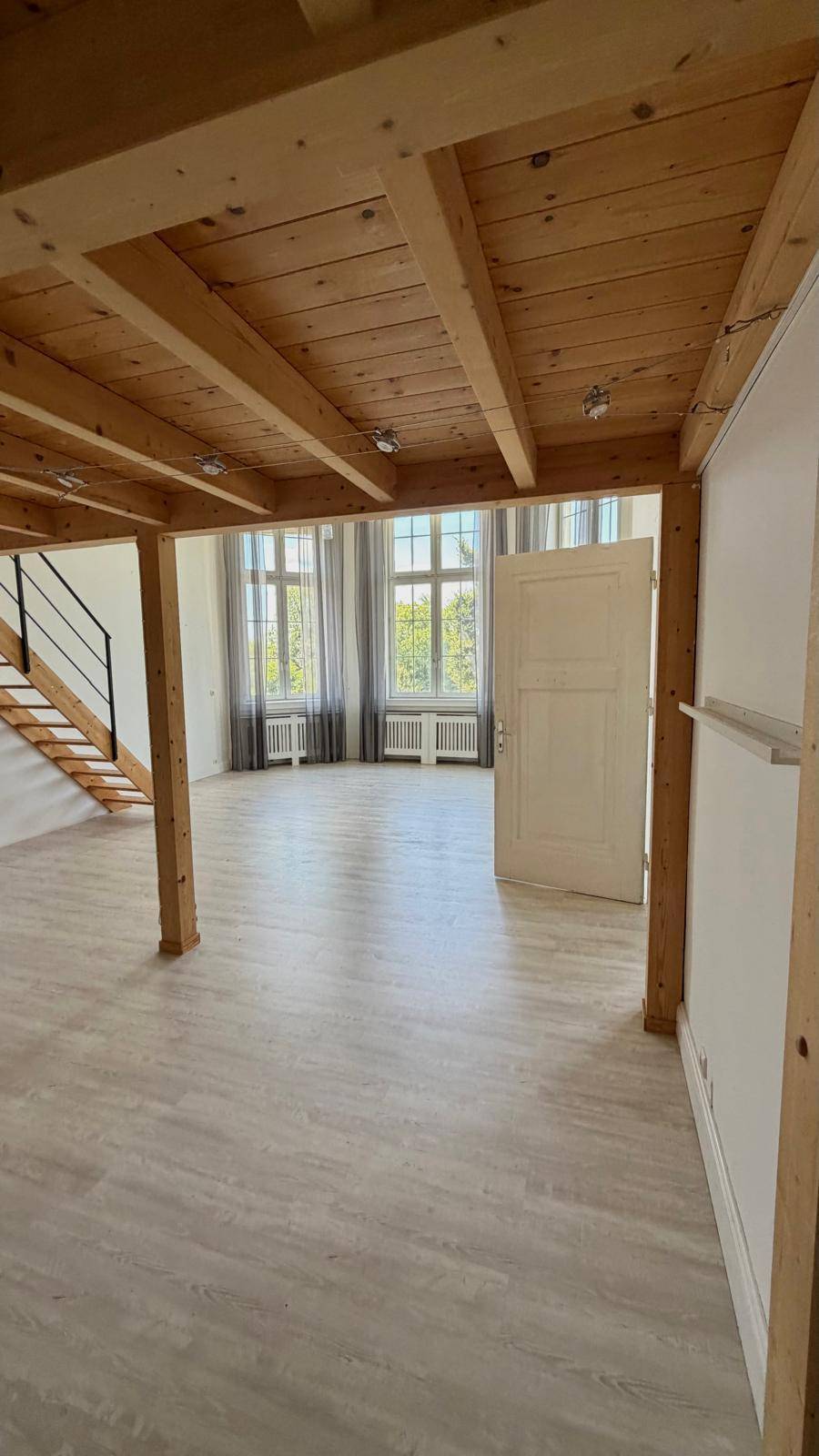 Offener Wohnbereich mit Holzbalkendecke und hellem Laminatboden, der zu einem weiteren Raum führt.