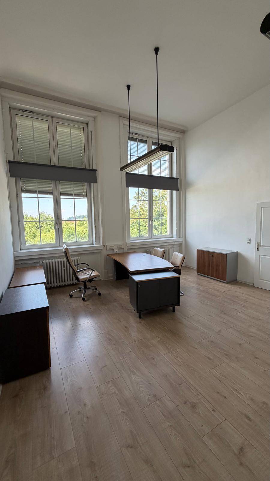 Weitläufiges Büro mit Laminatboden, einem großen Schreibtisch und zwei hohen Fenstern, die eine angenehme Arbeitsatmosphäre schaffen.