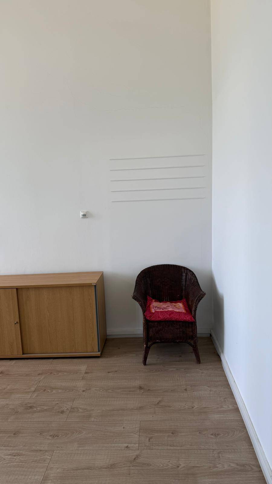 Ecke eines Büros mit weißer Wand, Holzschrank und einem Korbsessel mit rotem Kissen, schafft eine gemütliche Atmosphäre.