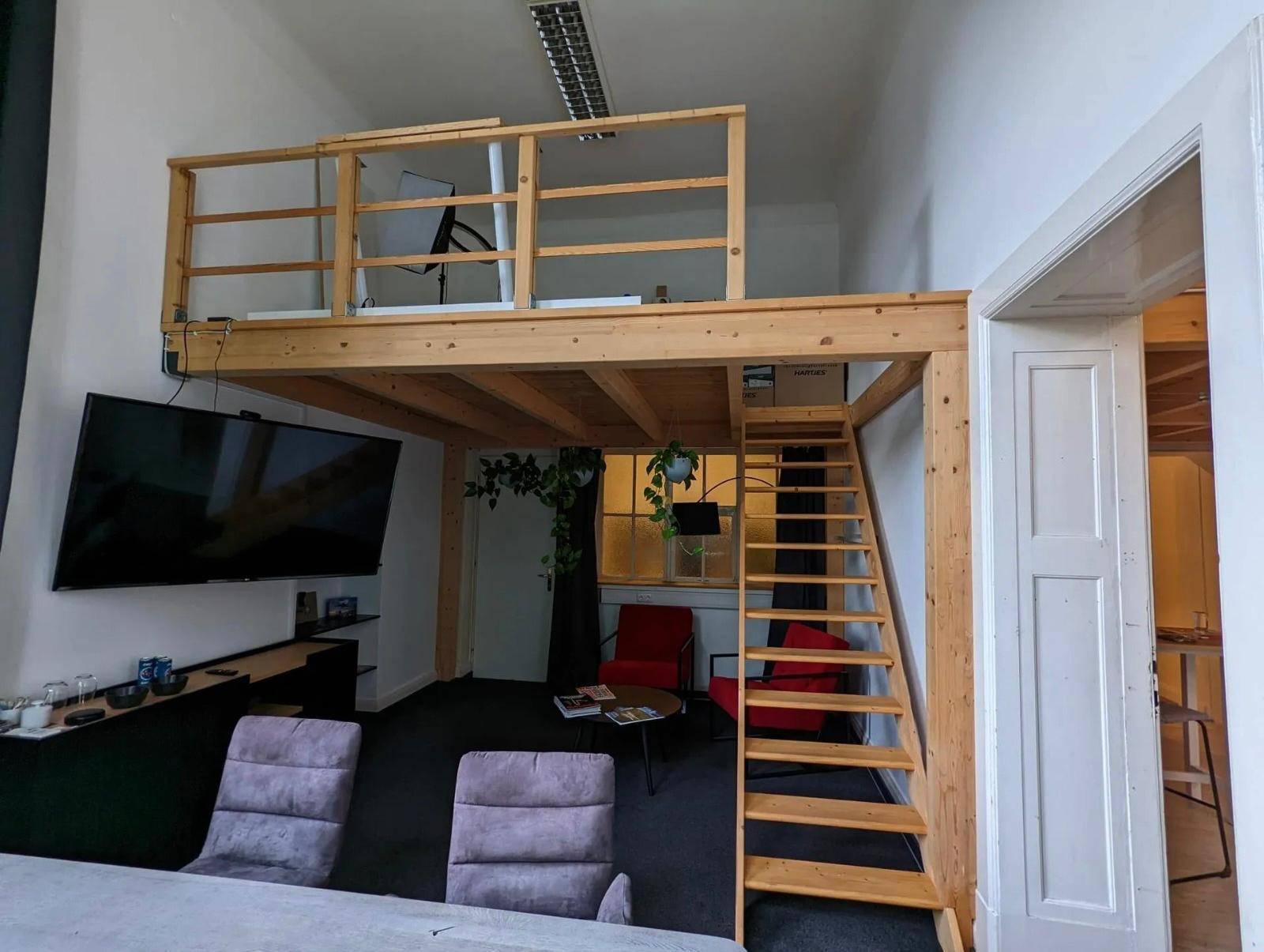 Innenansicht eines Büros mit Holz-Loft, Fernseher und roten Sesseln, die eine gemütliche Ecke bilden.