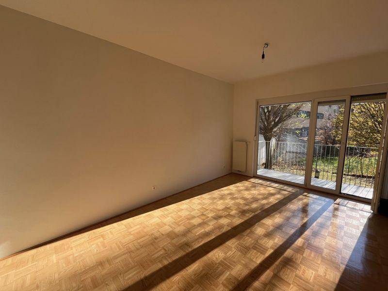 Geräumiges Zimmer mit Parkettboden und großem Fenster, das viel Tageslicht hereinlässt und einen Blick ins Grüne bietet.