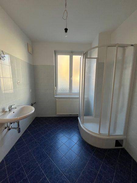 Badezimmer mit Duschkabine, Waschbecken und Fenster, in einem älteren Stil, der eine Renovierung benötigt.