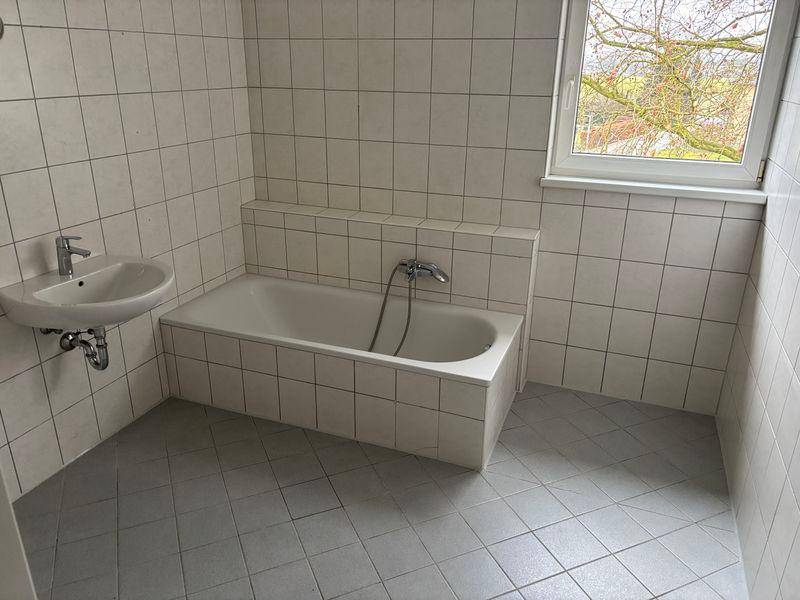 Badezimmer mit Badewanne, Waschbecken und Fenster, komplett gefliest in hellen Tönen.