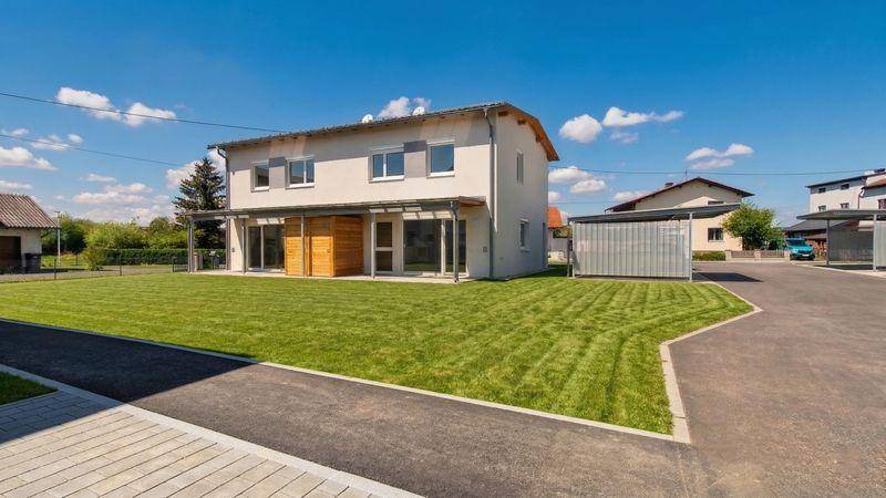 Modernes Einfamilienhaus mit heller Fassade, großem, gepflegtem Rasen und praktischen Carports unter strahlend blauem Himmel.