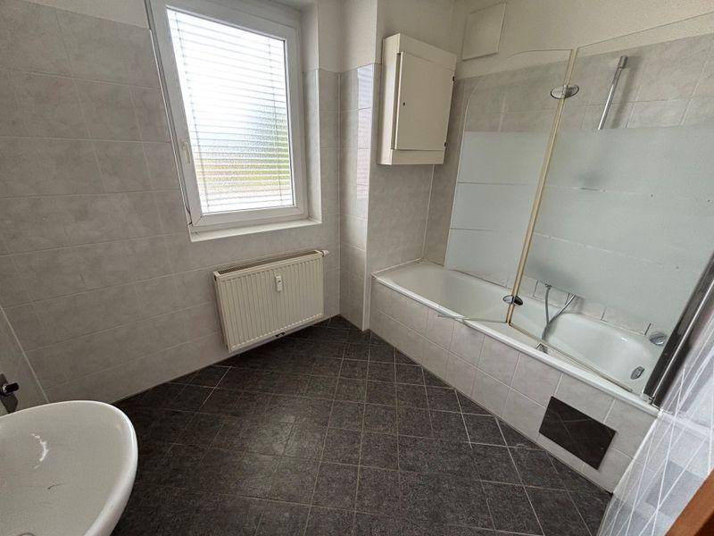 Badezimmer mit Badewanne, Waschbecken und Fenster, komplett gefliest in hellen Grautönen.