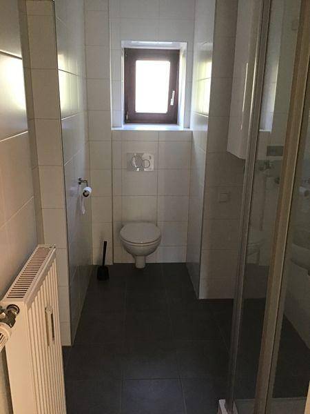 Schmales Badezimmer mit Toilette, Duschkabine, kleinem Fenster und weißen Fliesen.