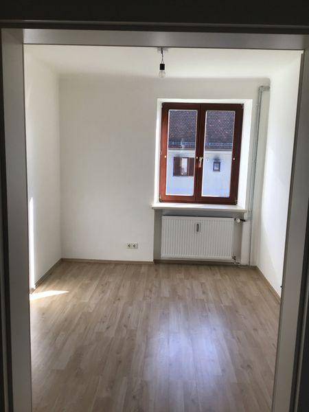 Heller, leerer Raum mit Laminatboden, einem Fenster mit Blick auf Dächer und einem Heizkörper.