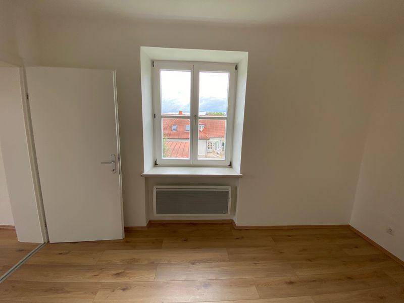 Schlafzimmer mit Laminatboden, Fenster und Blick auf die umliegenden Dächer und Berge.
