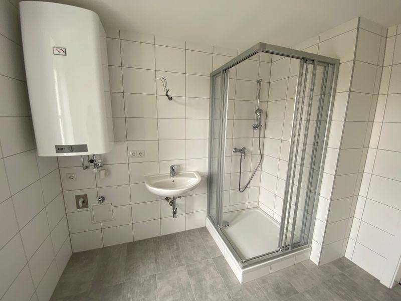 Badezimmerbereich mit Duschkabine, Waschbecken und Warmwasserboiler, gefliest in hellen Tönen.